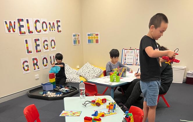 Children are playing LEGO around a large table with other various activities happening in the background, such as pouring out LEGO from a tub. 
